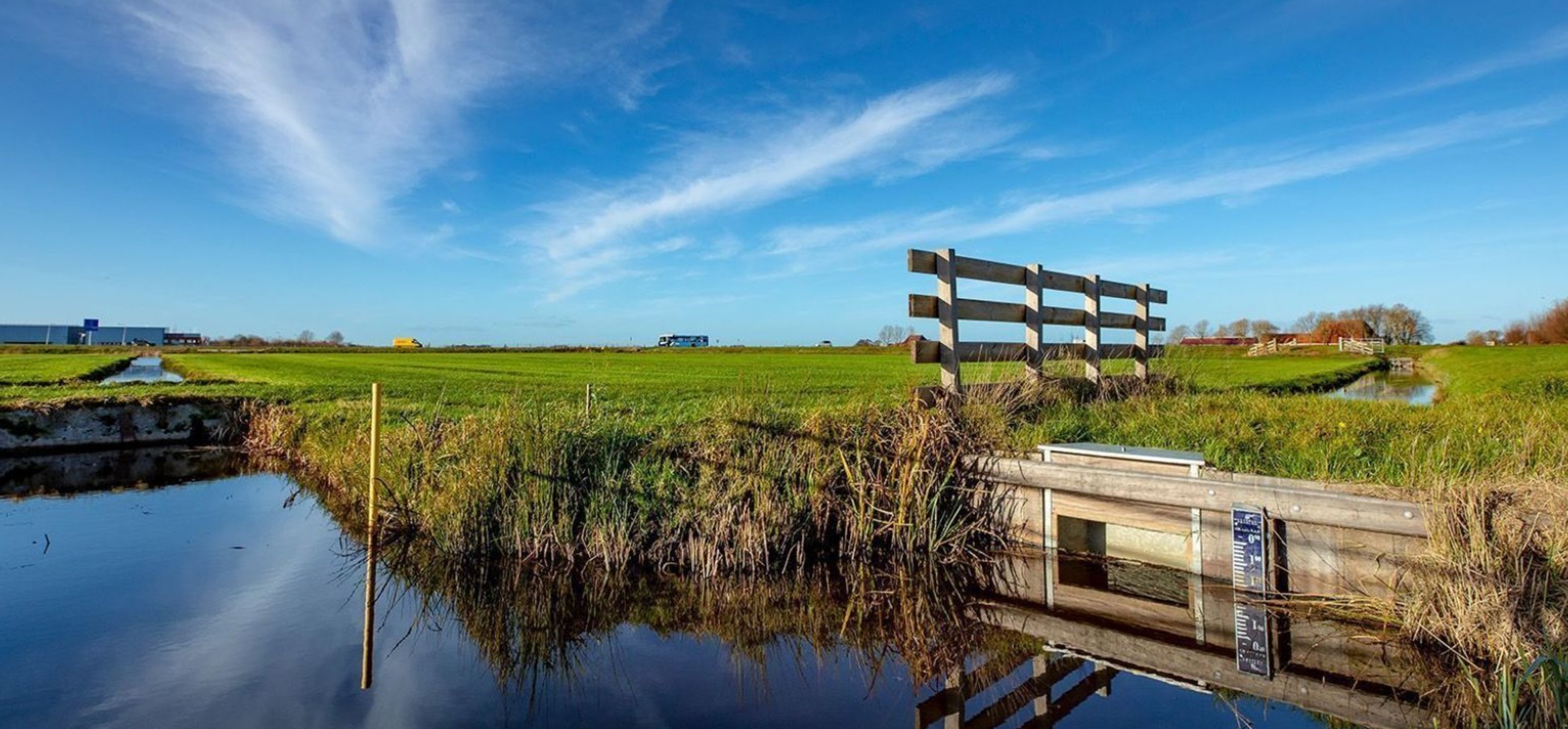 Foto van de oever van een weiland met houten beschoeiing en een waterpeil-meter